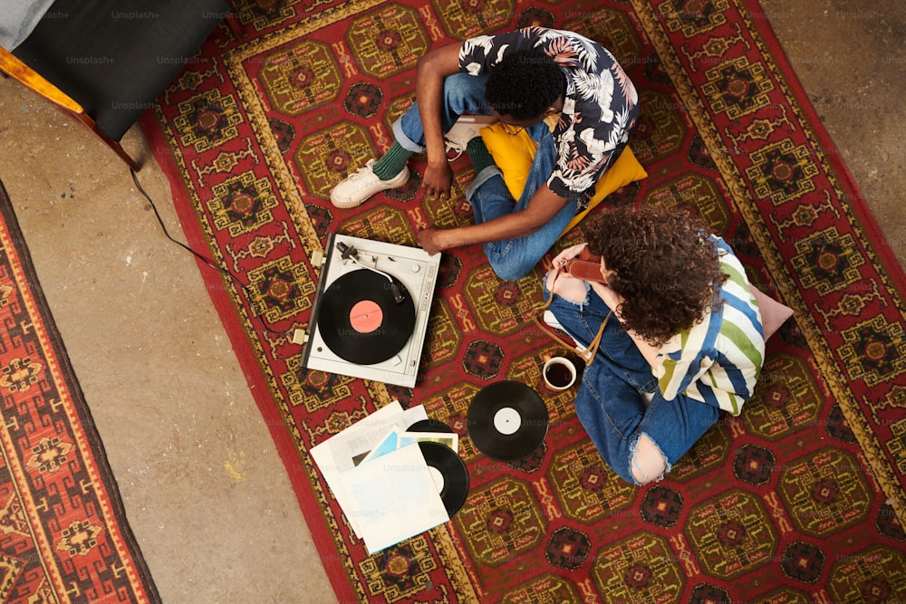 Above angle of two zoomers in stylish casualwear listening to vynil disks on record player while relaxing on red carpet in living room