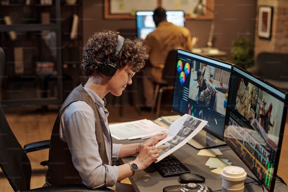 Redakteurin mit Kopfhörern, die das Skript an ihrem Arbeitsplatz untersucht, während sie Fotos am Computer bearbeitet