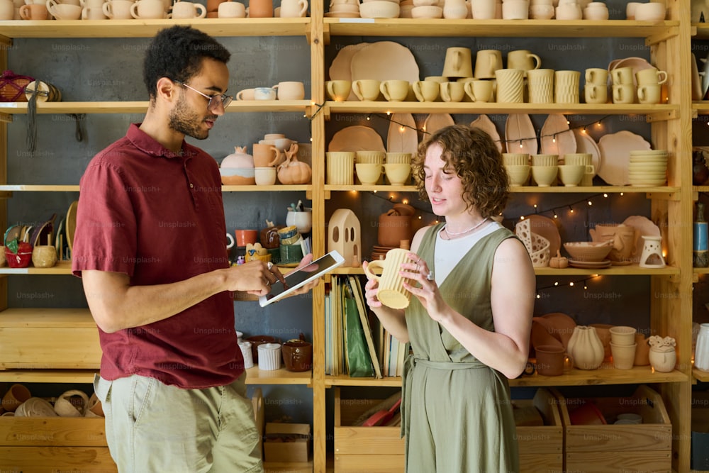 手作りの粘土マグカップを持つ若い女性が、その装飾やその他の特徴について同僚と展示に対して話し合う