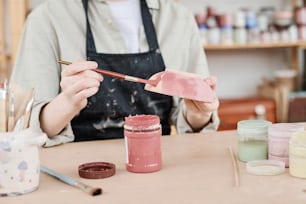 Giovane donna creativa in grembiule che tiene pennello e ciotola di argilla sopra il tavolo mentre colora la terracotta fatta a mano in marsala
