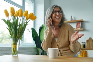 Glückliche Seniorin, die mit dem Handy telefoniert und gestikuliert, während sie sich zu Hause entspannt
