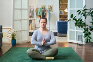 Junge Frau mit Behinderung sitzt in Lotuspose auf dem Boden eines geräumigen Wohnzimmers und schaut in die Kamera, während sie Yoga praktiziert