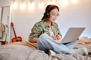 ベッドに足を組んで座りながらノートパソコンを使うアジアの10代の少女の全長のポートレート、コピー用スペース
