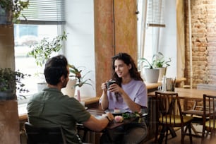 Happy brunette girl with cup of coffee and her boyfriend sitting by table in front of one another and discussing their interests in cafe