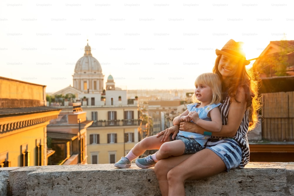 Mother and baby girl sitting on street overlooking rooftops of rome on sunset
