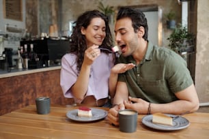 Young cheerful woman looking at her boyfriend eating piece of tasty cheesecake and having cappuccino during romantic date in cafe