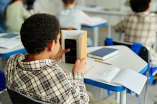 Vista trasera de un joven afroamericano escondiendo un teléfono inteligente en un libro y haciendo trampa en clase