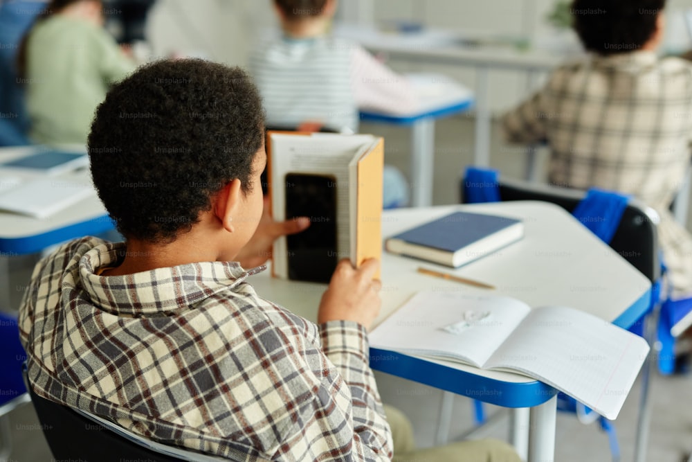 Back view at young African American kid hiding smartphone in book and cheating in class