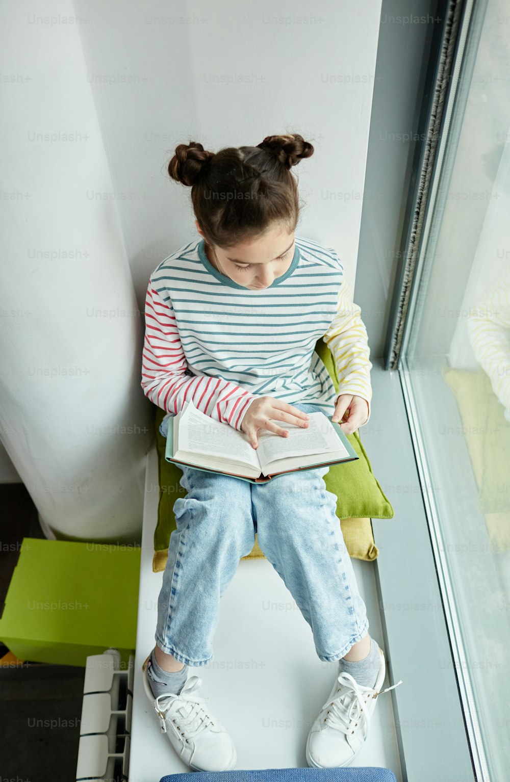 Vertical top view portrait of cute young girl reading book while sitting on window