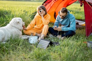 男性と女性は、自然を旅しながらキャンプ場で犬と一緒に座っています。ポータブルソーラーパネルでガジェットを充電します。旅行のための現代技術のコンセプト