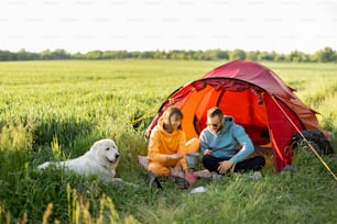 若い夫婦はテントの近くでピクニックをしながら、自然の中で犬と一緒に旅行します。夏の夜にグリーンフィールドでペットキャンプをする男女