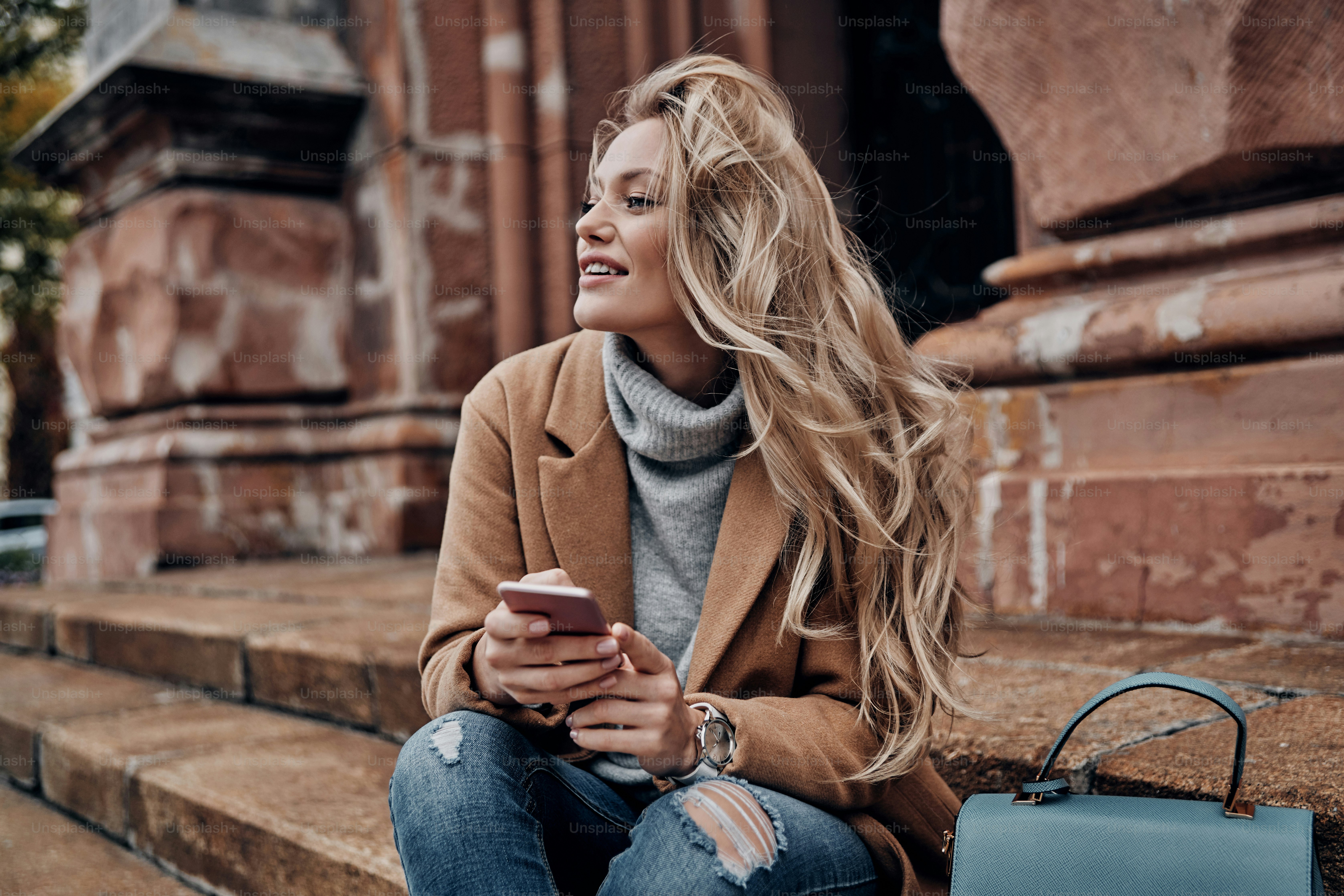 Beautiful young woman holding her smart phone and looking away with smile while sitting on stairs outdoors