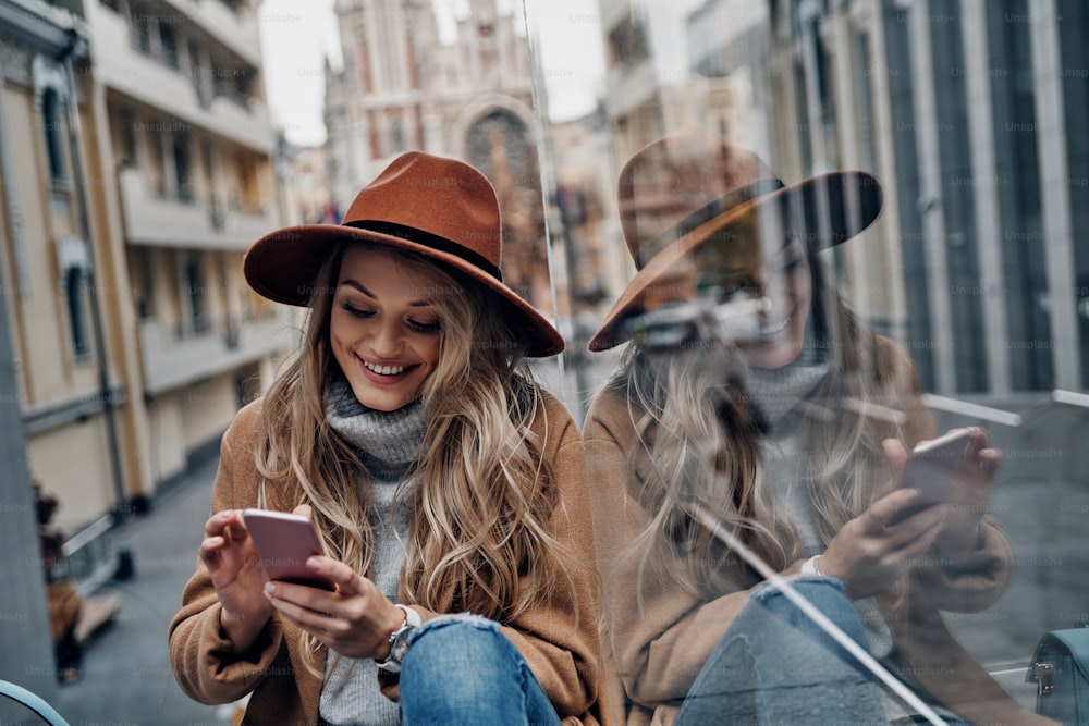 Attractive young woman in hat and coat using her smart phone while spending carefree time in the city