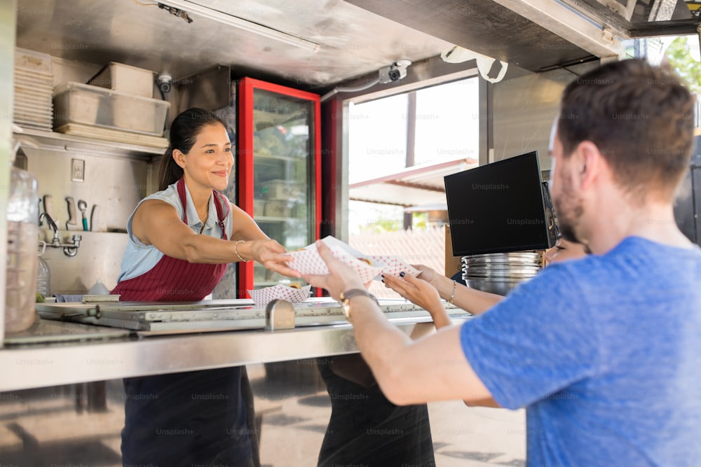 Giovane cuoca femminile carina che vende hot dog a un paio di clienti in uno stand gastronomico