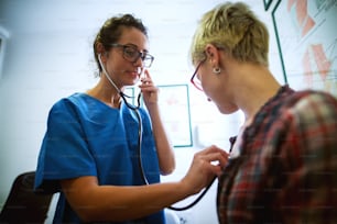 Portrait of professional middle aged nurse checking woman patient health condition with stethoscope in the office.