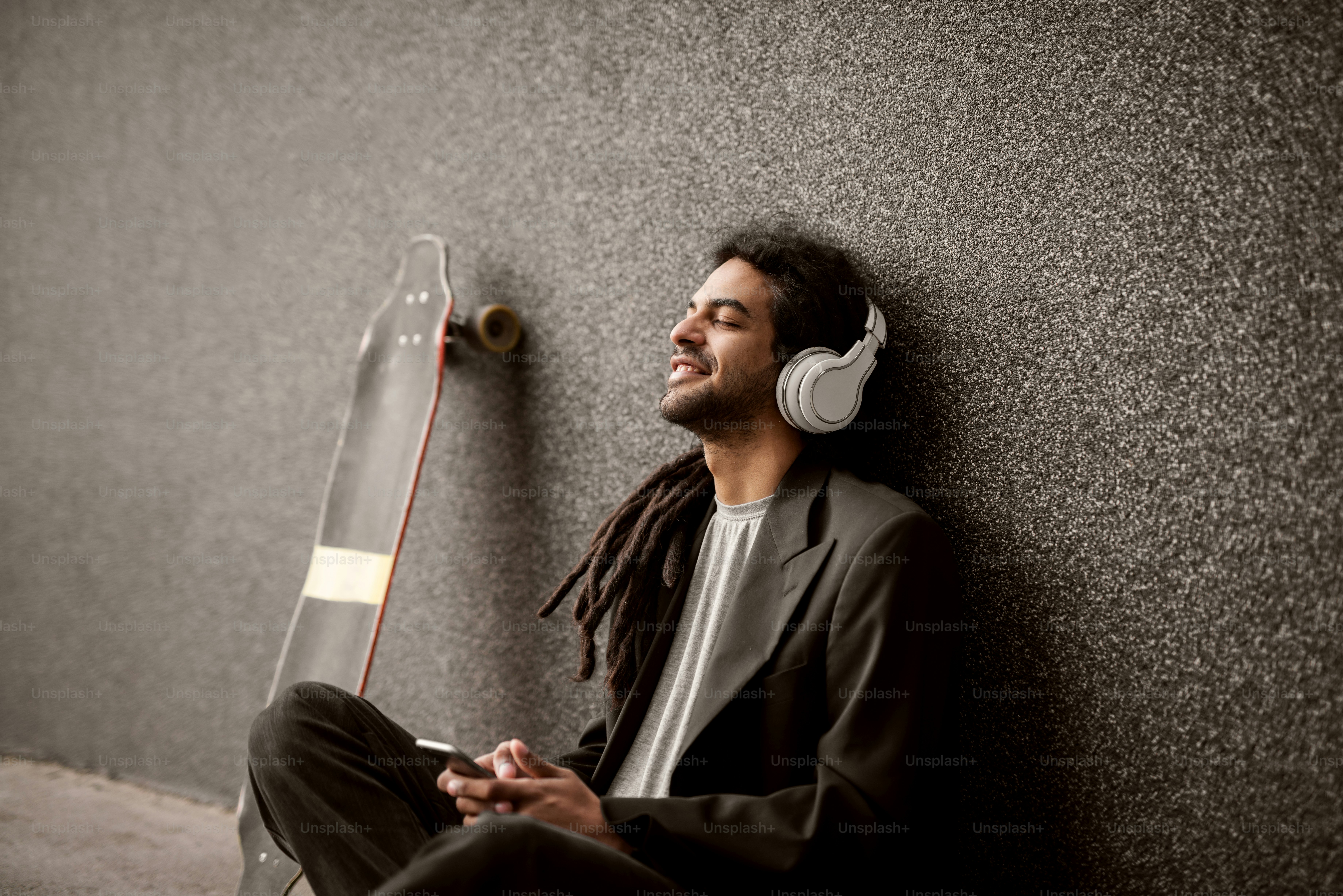 Stylish young dreadlocks hipster with headphones sitting leaning against the grey wall and skate near him listening music from a mobile.