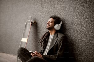 Stilvoller junger Dreadlock-Hipster mit Kopfhörern, der an die graue Wand gelehnt sitzt und in seiner Nähe skatet und Musik von einem Handy hört.