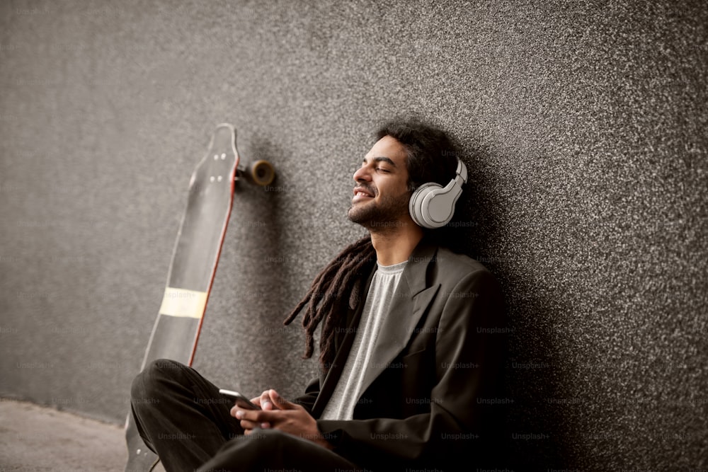 Élégant jeune hipster dreadlocks avec des écouteurs assis appuyé contre le mur gris et patiner près de lui en écoutant de la musique depuis un mobile.