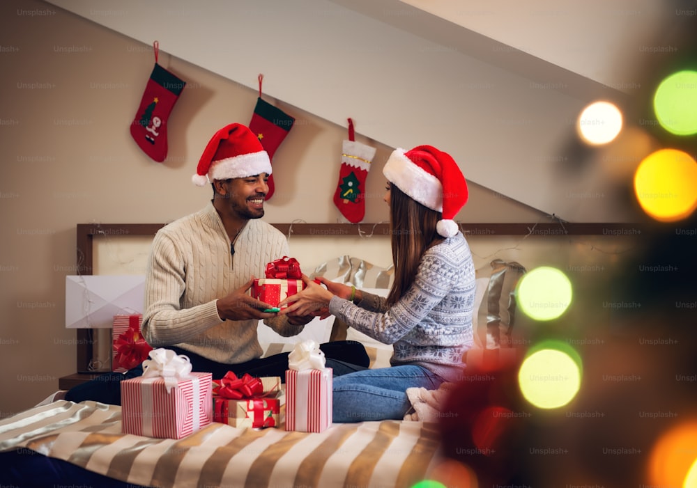 Feliz pareja joven y guapa emocionada dándose regalos de Navidad mientras están sentados en la cama con suéteres y sombreros de Papá Noel.
