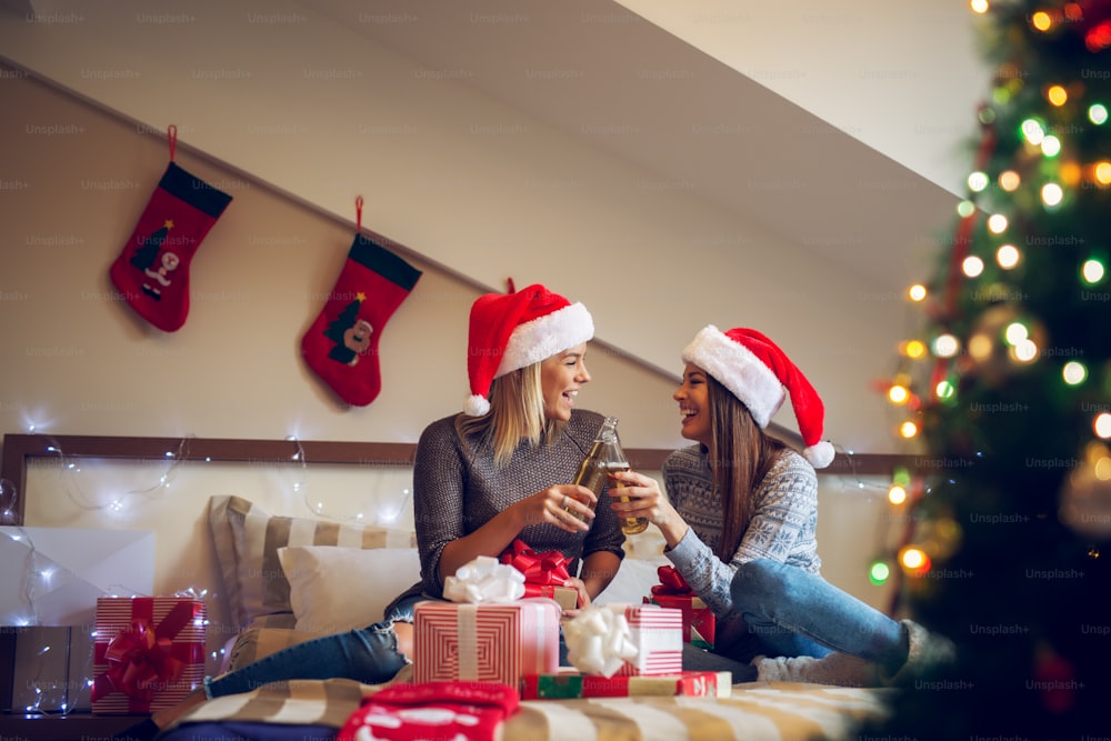 サンタの帽子をかぶった2人の陽気なガールフレンドがクリスマス休暇にベッドに座り、ビールで歓声を上げ、プレゼントや装飾品が周りにあります。