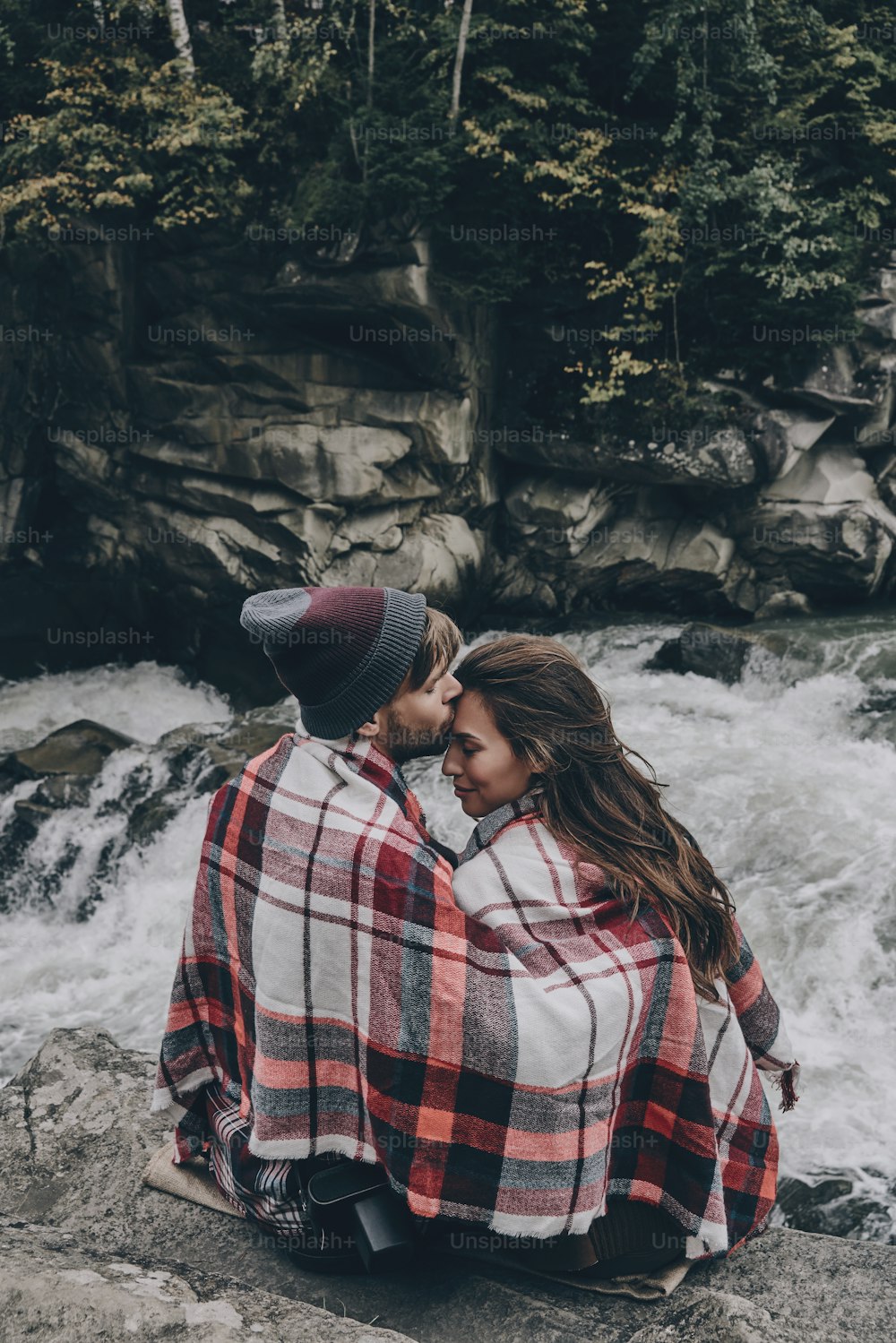 Joven guapo besando a una joven atractiva mujer mientras está sentado en las rocas con el río debajo