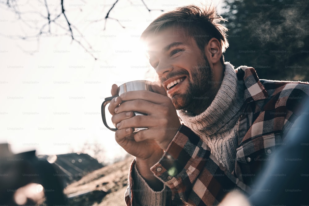따뜻한 옷을 입은 잘생긴 청년이 모닝 커피를 마시고 산에서 캠핑을 하며 웃고 있다