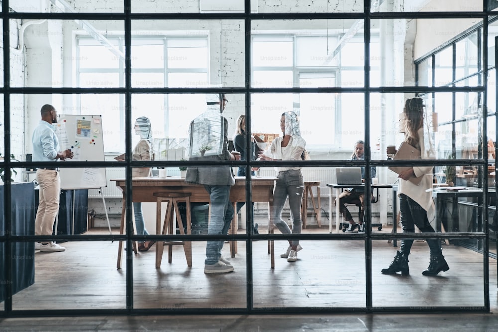 Gran grupo de jóvenes modernos que trabajan y se comunican juntos mientras están parados detrás de la pared de vidrio en la sala de juntas