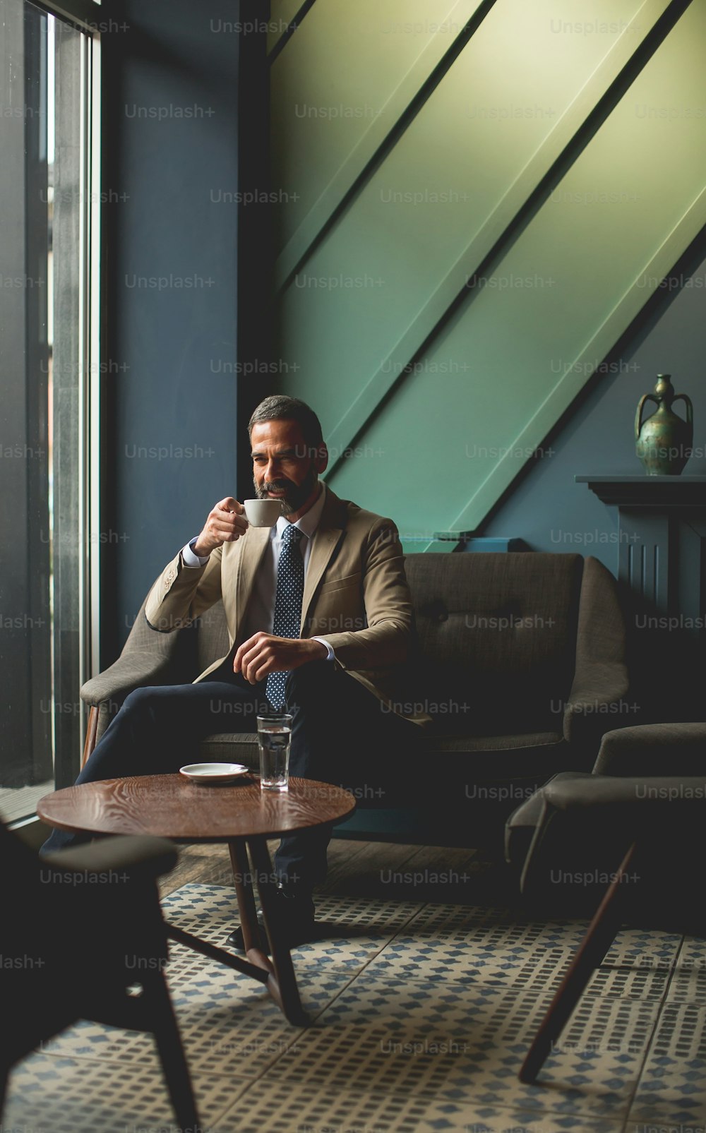 Handsome elegant serious businessman sitting in armchair and holding cup of espresso in cafe