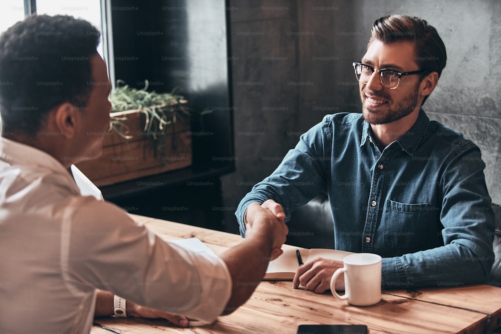 Young modern men in smart casual wear shaking hands and smiling while spending time in the office