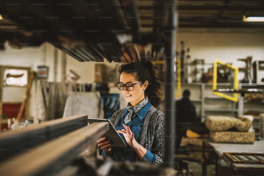 Close up view of hardworking focused professional motivated business woman holding a tablet next to the shelf with metal pipes in the fabric workshop.