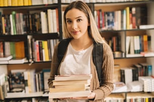 Porträt eines jungen, schönen, nerdigen blonden Studentenmädchens, das einen Bücherstapel in der sonnigen Bibliothek hält.