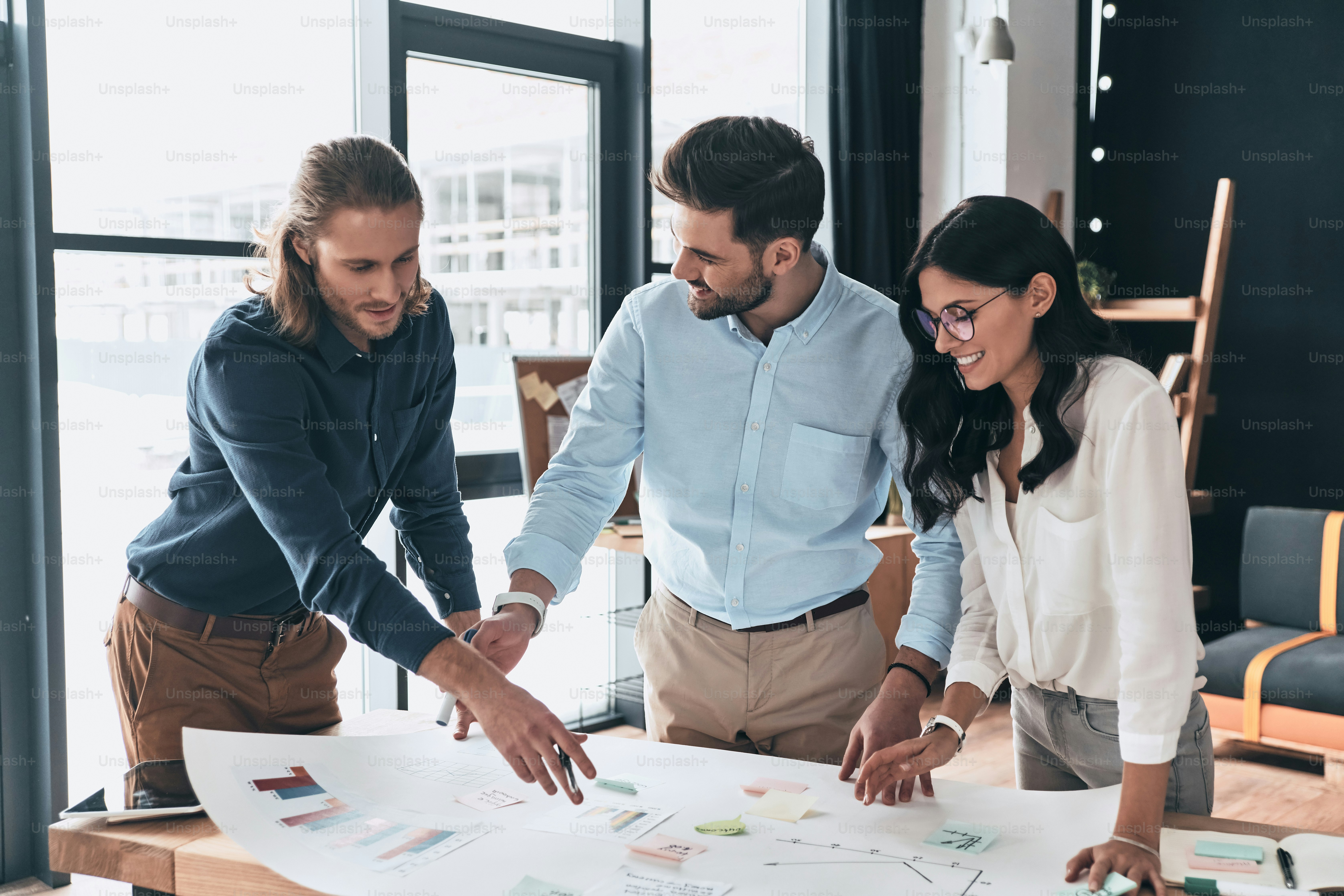 Group of young confident business people discussing something and smiling while looking at blueprint in the office