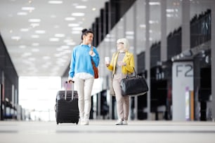 Dos chicas con equipaje caminando por el edificio del aeropuerto antes o después del check-in y conversando