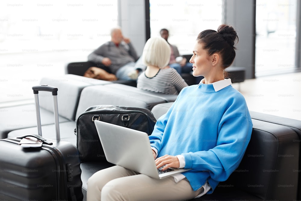 青いプルオーバーを着た若い実業家が空港ラウンジに座り、フライトを待っている間にネットで働く