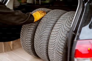 Hard working mechanic in orange gloves is putting car wheels into a car trunk carefully.