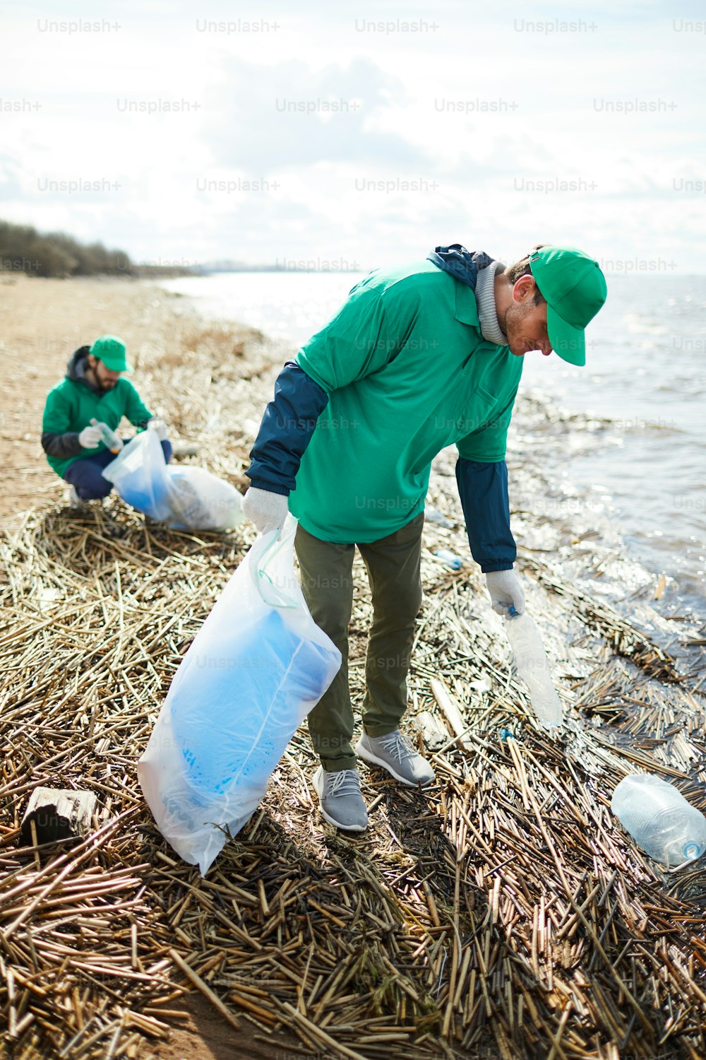 Young greenpeacers in uniform walking along waterside and picking up litter for utilization