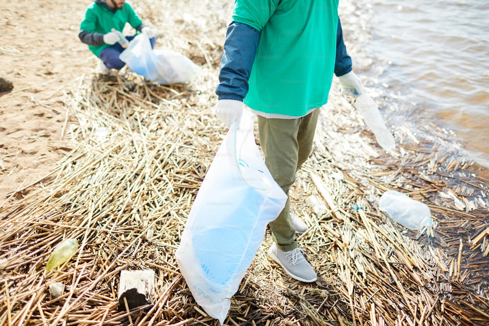 Dois humanos de uniforme verde com sacos limpando área ribeirinha de garrafas plásticas velhas