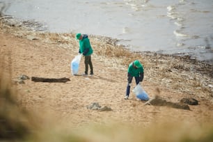 Two youn men in green uniform cleaning river bank from litter and putting it into sacks