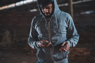 Portrait of hipster afro-american young handsome man relaxing outside on the street at night with earphones and cigarette while choosing music on a mobile.