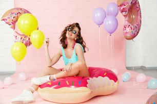 Moda de verano. Hermosa mujer en traje de baño de moda. Feliz modelo femenina sonriente con cuerpo sexy en elegante traje de baño sentado en dona inflable sobre fondo rosa. Alta resolución.
