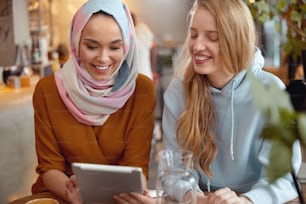 Women. Friends Meeting In Cafe. Smiling Girls Looking At Tablet Screen. Different Ethnicity Female Having Fun In Bistro.