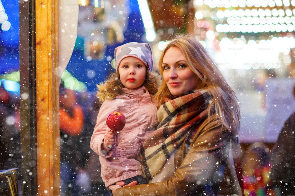 Beautiful woman and little kid girl eating crystallized sugared apple on German Christmas market. Happy family, mother and daughter in winter clothes with lights on background. Tradition, holiday concept