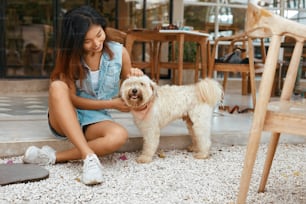 Mujer con perro en la terraza. Hermosa modelo asiática en ropa de moda abraza a la mascota en el patio en un café que admite perros. Mujer feliz en traje de jeans jugando con cachorro y disfrutando de las vacaciones de verano.