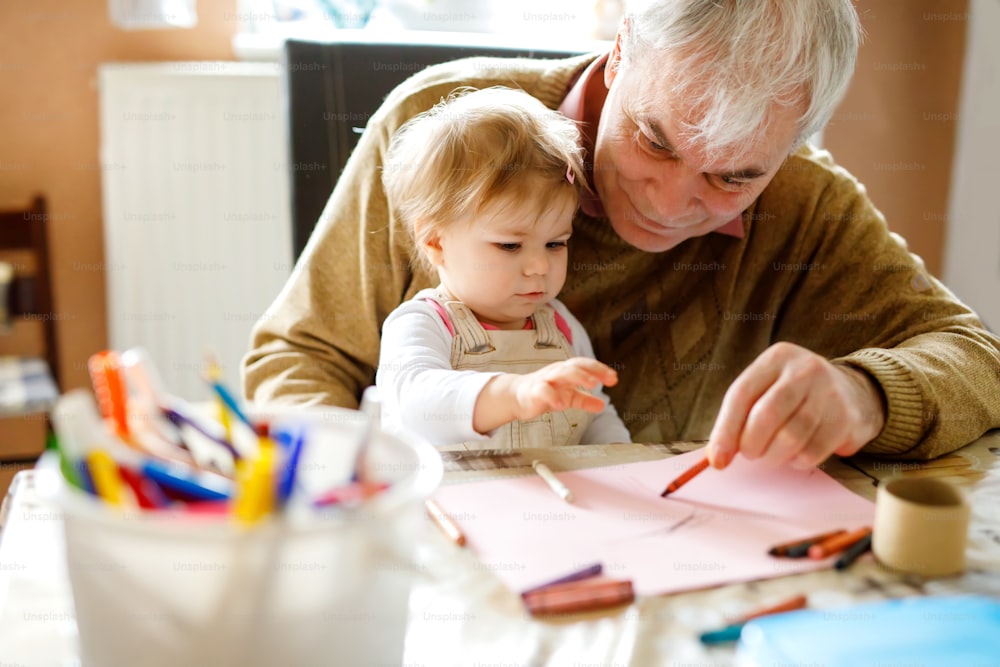 Cute little baby toddler girl and handsome senior grandfather painting with colorful pencils at home. Grandchild and man having fun together. Family and generation in love.