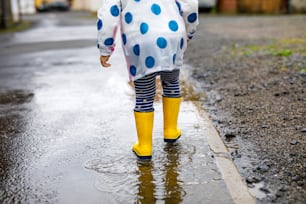 黄色いレインブーツを履き、雨の曇りの日にみぞれの間を歩く小さな幼児の女の子の接写。カラフルな服を着たかわいい子供が水たまりに飛び込み、水しぶきを上げ、野外活動を行う。
