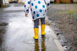 Close-up of little toddler girl wearing yellow rain boots and walking during sleet on rainy cloudy day. Cute child in colorful clothes jumping into puddle, splashing with water, outdoor activity.