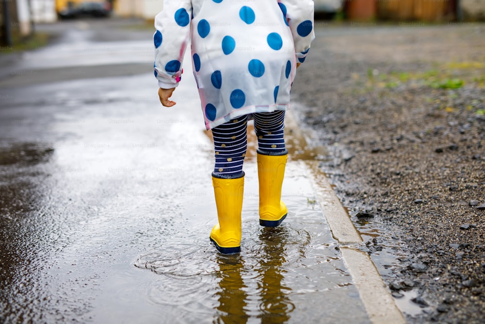 Close-up of little toddler girl wearing yellow rain boots and walking during sleet on rainy cloudy day. Cute child in colorful clothes jumping into puddle, splashing with water, outdoor activity.