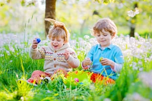 Two little kids boys and friends in Easter bunny ears during traditional egg hunt in spring garden, outdoors. Siblings having fun with finding colorful eggs. Old christian and catholoc tradition