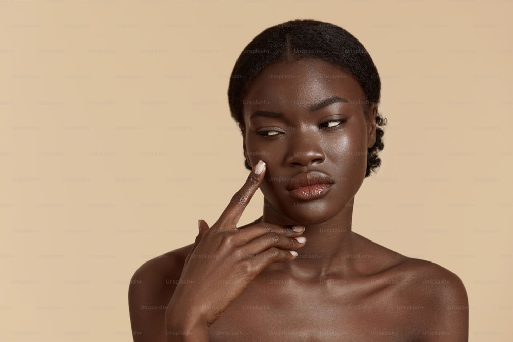 Portrait close up of beautiful african girl. Thoughtful young woman touch her clean face. Concept of face skin care. Isolated on beige background. Studio shoot