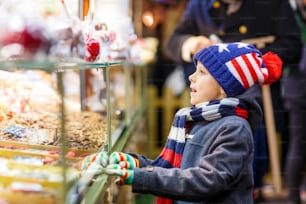 진저브레드와 견과류를 곁들인 달콤한 스탠드 근처에 있는 작은 귀여운 소년. 독일의 크리스마스 시장에서 행복한 아이. 크리스마스에 가족을 위한 전통적인 레저. 휴일, 축하, 전통, 어린 시절.
